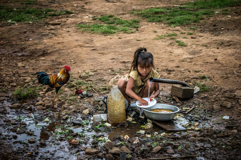 An asian child washing dishes in an outdoor setting, with a rooster beside her.